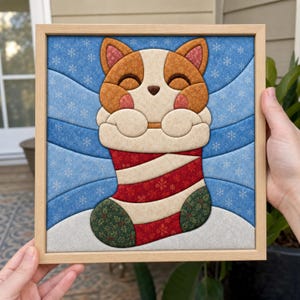 Peut inclure: Une œuvre d'art en tissu encadrée représentant un chat stylisé au pelage orange et crème sortant d'une chaussette de Noël rayée rouge et blanche. Le fond est en tissu bleu à motifs de flocons blancs, le tout dans un cadre en bois clair.