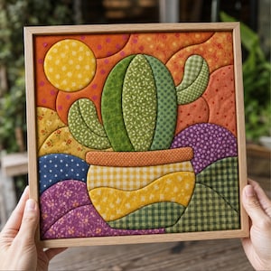 May include: A square fabric wall art piece in a light wood frame featuring a quilted cactus in a pot. The cactus is green with various patterns, set against a background of orange, yellow, purple, and blue fabric shapes with floral and gingham prints.