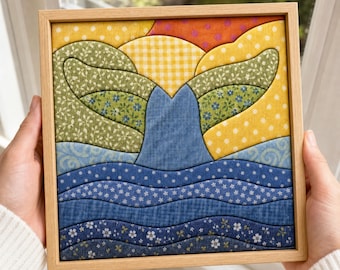 May include: A square fabric wall art piece in a light wood frame featuring a quilted whale tail design. The composition uses various patterned fabrics in shades of blue, green, yellow, and orange to depict a whale tail emerging from wavy ocean water.