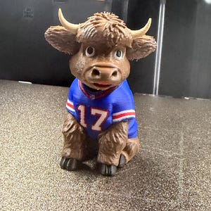 May include: A brown Highland cow figurine with textured fur and light-colored horns, wearing a blue sports jersey with the number 17 printed in white on the front, accented with red and white stripes on the sleeves and collar, sitting in an upright position.