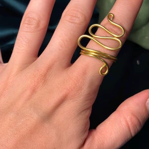 May include: A gold-coloured wire ring featuring a continuous, winding design that wraps around the finger. The metal wire forms a series of interconnected loops and curves, creating an open, layered structure that sits prominently on the finger.