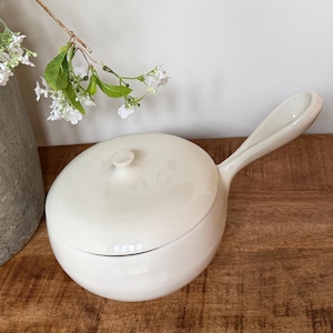 May include: A cream-colored ceramic saucepan with a matching lid and a single elongated handle. The pot features a smooth, glossy finish and a rounded body shape. The lid includes a small, centered knob for lifting, and the handle extends outward.