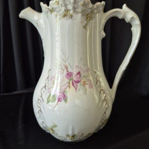 May include: A white porcelain coffee pot featuring a delicate floral pattern with pink and purple flowers, green leaves, and gold trim accents. The vessel has a scalloped rim, a curved handle, a spout, and a lid with a decorative finial handle.
