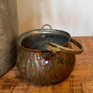 May include: A small, round copper pot featuring an embossed leaf pattern on its exterior. The vessel includes a metal swing handle attached with rivets and a dark, weathered patina throughout the surface, showing signs of age and oxidation.