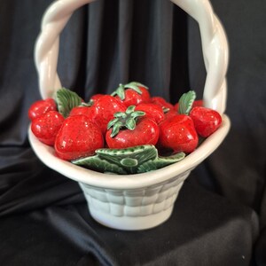 May include: A white ceramic basket with a braided handle filled with glossy red strawberries and green leaves. The basket features a woven texture on the exterior, while the strawberries are sculpted with realistic details and vibrant red glaze.