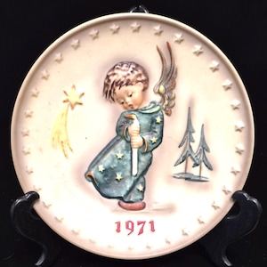 May include: A round ceramic decorative plate featuring a raised relief of an angel in a dark teal robe holding a white candle. The plate has a cream background with a border of stars, a yellow shooting star, two pine trees, and the year 1971 in red.