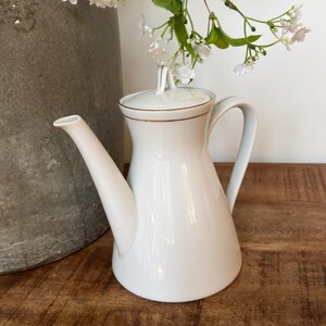May include: A white porcelain teapot with a smooth, tapered body, a curved handle, and a matching lid featuring a small finial. The rim of the lid and the top edge of the teapot are accented with a thin, metallic gold line, providing a subtle contrast.
