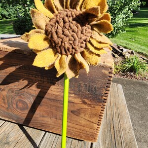 May include: A handcrafted sunflower decoration featuring layered mustard yellow felt petals and a textured dark brown center. The flower is attached to a bright lime green stem, creating a vibrant botanical accent piece with a distinct fabric texture.