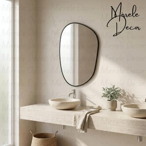 May include: An irregular, organic-shaped wall mirror with a thin black frame hangs above a light-colored stone vanity. The vanity features two round stone vessel sinks, a silver faucet, a small potted plant, and a folded beige cloth.