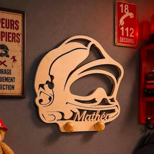Può includere: Un appendiabiti da parete in legno chiaro a forma di casco da vigile del fuoco con un design intagliato. Il nome Mathéo è inciso sulla parte anteriore e due pomelli rotondi in legno sono fissati in basso per appendere oggetti.