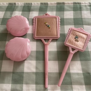 以下が含まれることがあります： 2つの貝殻型の蓋付き容器と、正方形のフレームが付いた2つの手鏡を含む、4つのライトピンクの化粧用アクセサリーセット。各鏡には、小さな花の装飾が施されたベージュのテクスチャ加工された中央パネルが付いています。