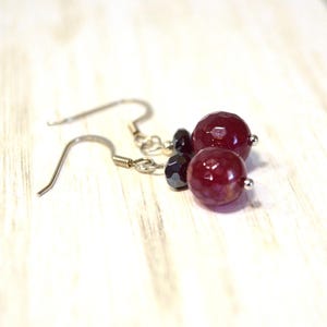 May include: A pair of dangle earrings featuring faceted, deep red beads and smaller black beads. The earrings have silver-colored ear wires and are displayed on a light-colored wooden surface. The earrings have a simple, elegant design.