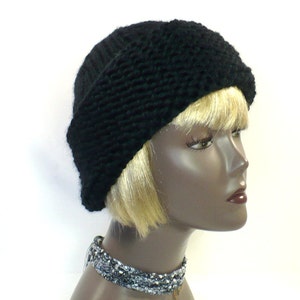 May include: A solid black knit beanie featuring a thick, textured brim and a smooth, ribbed crown. The hat is displayed on a mannequin head with blonde hair, accompanied by a multi-strand, metallic silver and grey beaded choker necklace.