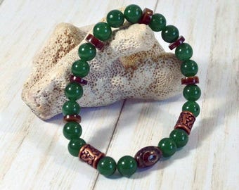 May include: A beaded bracelet featuring round, polished green beads interspersed with dark brown, flat disc-shaped spacers and two cylindrical copper-toned metal beads with intricate embossed patterns, plus one oval brown bead with a central eye design.