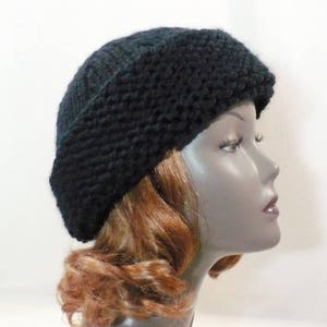 May include: A black knit beanie hat with a thick, textured garter stitch pattern and a wide, folded brim. The hat is crafted from a chunky yarn, providing a dense and warm appearance. It is displayed on a mannequin head with auburn hair.