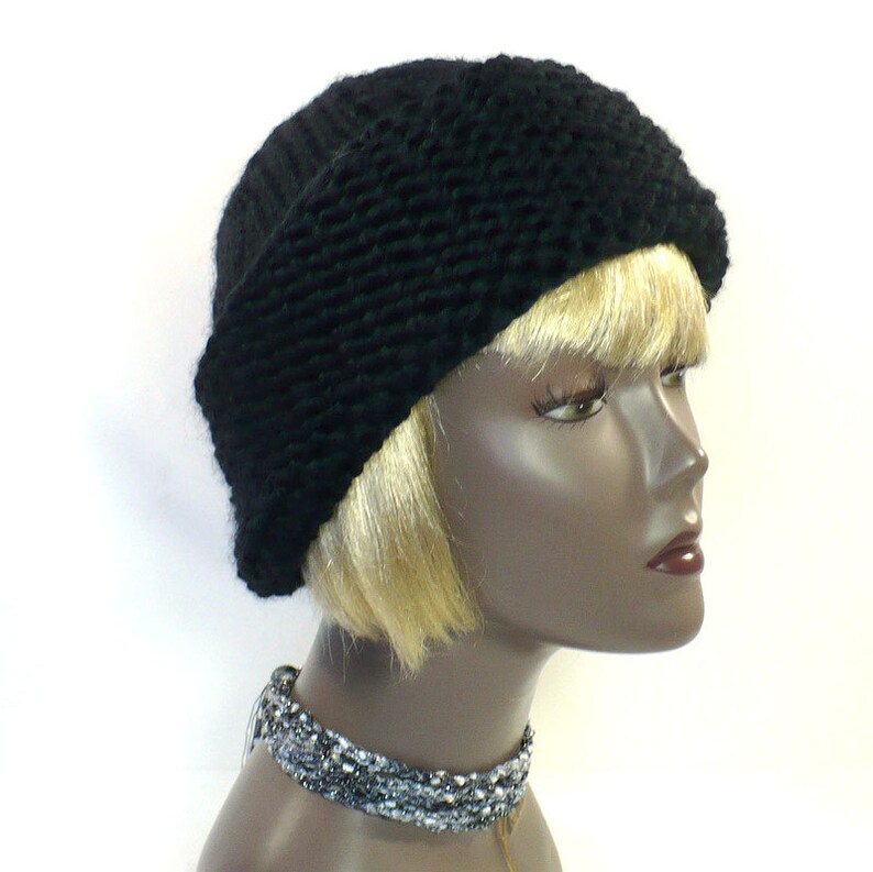 May include: A solid black knit beanie featuring a thick, textured brim and a smooth, ribbed crown. The hat is displayed on a mannequin head with blonde hair, accompanied by a multi-strand, metallic silver and grey beaded choker necklace.