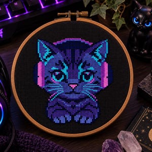 May include: A circular wooden embroidery hoop containing a cross-stitch pattern of a cat wearing pink headphones. The cat is rendered in shades of dark blue, purple, and bright cyan against a solid black background, featuring large eyes and whiskers.