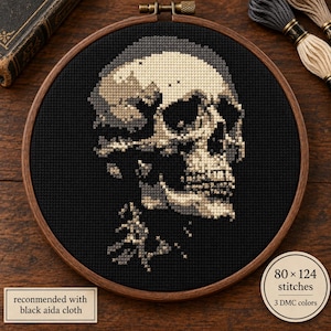 May include: A cross-stitch pattern of a human skull rendered in shades of cream, gray, and black on black fabric, secured in a circular wooden embroidery hoop. The design includes text stating 80 x 124 stitches and recommended with black aida cloth.