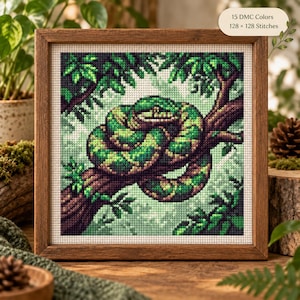 May include: A square cross-stitch pattern in a wooden frame depicting a coiled green and brown snake resting on a tree branch. The design features 15 DMC colors and a 128 x 128 stitch count. The background shows foliage in shades of green.