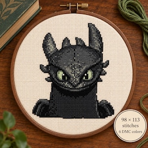Può includere: Un ricamo a punto croce in un telaio circolare in legno raffigurante un drago nero con grandi occhi verdi su un tessuto color crema. Una piccola etichetta circolare sul lato mostra il testo 98 x 113 stitches e 6 DMC colors.