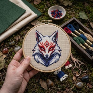 May include: A circular wooden embroidery hoop containing a cross-stitch pattern of a stylized fox head. The fox features white fur with intricate red and dark blue geometric markings around the eyes and ears, set against a light beige fabric background.