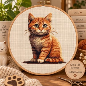 Könnte beinhalten: Ein runder Holzstickrahmen mit einem Kreuzstichmuster einer sitzenden orangefarbenen getigerten Katze mit grünen Augen. Ein kleines rundes Etikett in der Ecke zeigt den Text 128 x 128 stitches und 16 DMC colors mit einem Pfotenabdruck-Symbol.