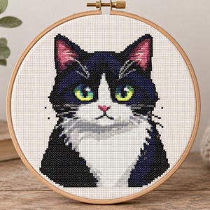 May include: A cross-stitch embroidery piece in a round wooden hoop featuring a portrait of a black and white tuxedo cat. The cat has bright green eyes, a small pink nose, and pointed ears with pink inner details, set against a white fabric background.