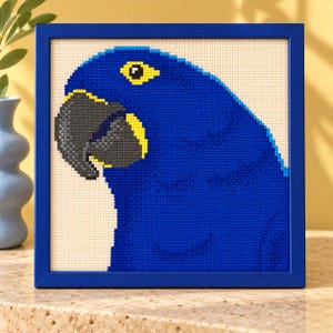 May include: A square framed diamond painting featuring a pixelated blue macaw parrot with a dark gray beak and a bright yellow eye patch, set against a cream-colored background within a solid blue frame.