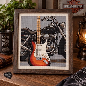May include: A framed cross-stitch embroidery featuring a sunburst electric guitar layered over a grayscale motorcycle. The guitar has a white pickguard and a light-colored neck. The artwork is enclosed in a dark brown wooden frame with a textured finish.