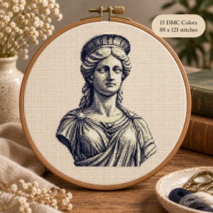Op de afbeelding: Een ronde houten borduurring met een kruissteekpatroon van een klassieke vrouwenbuste in blauw- en wittinten. De stof bevat de tekst 15 DMC Colors en 88 x 121 stitches. Het ontwerp toont een figuur met gevlochten haar.