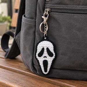 May include: A crochet keychain featuring a white and black ghost mask design with a long, open mouth and two oval eyes. The textile charm is attached to a silver-toned metal lobster clasp and a circular split ring, hanging from a dark gray backpack.