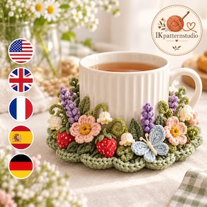 May include: A white ceramic mug sits inside a round, green crocheted coaster decorated with three-dimensional crocheted flowers, red strawberries, white lily of the valley, and a light blue butterfly. The logo IKpatternstudio is visible.