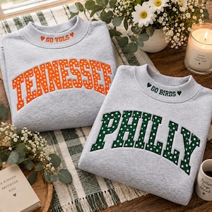 Op de afbeelding: Twee lichtgrijze sweatshirts met ronde hals. Eén heeft oranje letters met witte stippen die TENNESSEE vormen en de tekst GO VOLS met twee hartjes. De andere heeft groene letters met witte stippen die PHILLY vormen en de tekst GO BIRDS met twee hartjes.