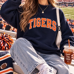 May include: A navy blue long-sleeve sweatshirt featuring the word TIGERS in orange block letters with white polka dots across the chest. The sweatshirt is paired with light blue denim jeans, a white shoulder bag, and a gold necklace.