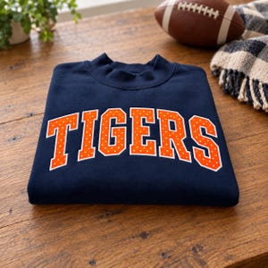 May include: A navy blue crewneck sweatshirt featuring the word TIGERS in large, arched, orange block letters with a white polka dot pattern. The text is centered on the chest of the garment, which is folded neatly on a wooden surface.