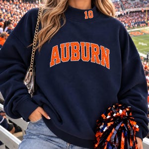 Puede incluir: Una sudadera azul marino de cuello alto con la palabra AUBURN en letras grandes de color naranja con un patrón de puntos blancos en el pecho y el número 10 en naranja en el cuello. La prenda tiene mangas largas y un diseño de corte holgado.