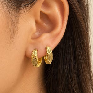 May include: A pair of small gold hoop earrings with a textured, geometric, faceted surface. The earrings have a metallic yellow finish and a thick, chunky design that sits close to the earlobe, showcasing a distinct angular pattern on the outer edge.