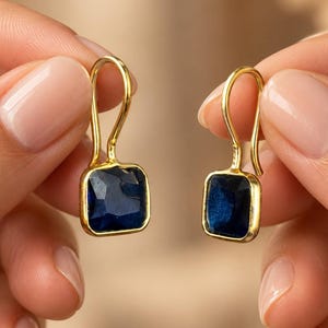 Puede incluir: Un par de pendientes colgantes de tono dorado con piedras facetadas de color azul intenso de corte cuadrado engastadas en marcos de metal pulido. Los pendientes cuelgan de ganchos curvos, mostrando un diseño sencillo con un contraste azul.