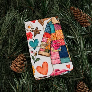 May include: A rectangular gift wrapped in white paper featuring a colorful patchwork jacket illustration, red and teal hearts, white daisies, and brown star-shaped accents. The package is tied with thin brown twine and sits among pine needles.