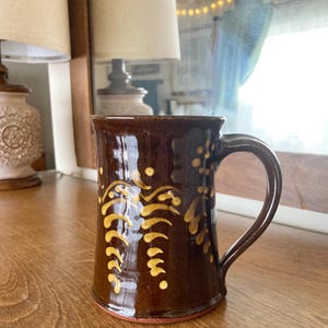 May include: A brown ceramic mug with a handle and a decorative yellow floral design. The mug has a dark brown glaze and a red base. The mug is sitting on a wooden surface.