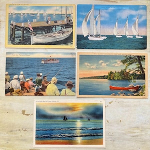 May include: A collection of vintage postcards featuring nautical themes. The postcards depict sailboats, fishing boats, and coastal landscapes. One postcard shows a pier with a boat and an American flag. Another shows a red canoe on a lake.