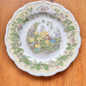 May include: A decorative plate with a scalloped edge and a cream-colored border. The plate features a detailed illustration of a spring scene with flowers, a small house, and the text "Brambly Hedge Spring". The plate is on a wooden surface.