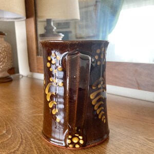 May include: A brown ceramic mug with a handle, decorated with yellow floral patterns. The mug has a glossy finish and a wide base. The design includes a cluster of yellow dots at the base of the handle.