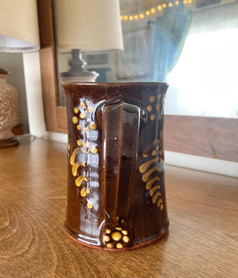 May include: A brown ceramic mug with a handle, decorated with yellow floral patterns. The mug has a glossy finish and a wide base. The design includes a cluster of yellow dots at the base of the handle.