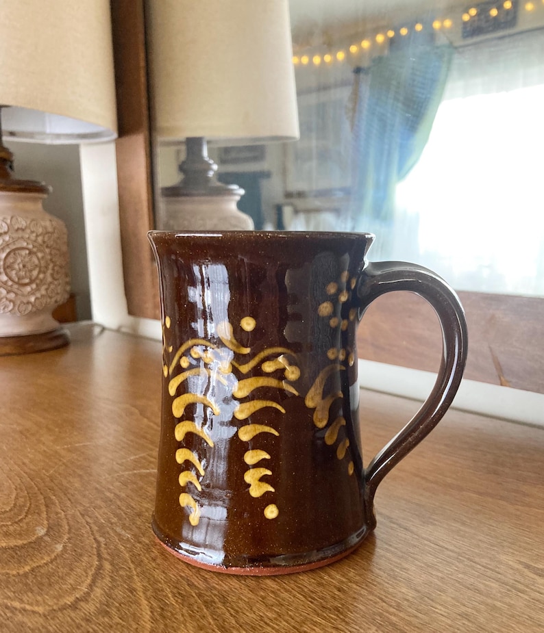May include: A brown ceramic mug with a handle and a yellow floral design. The mug has a dark brown glaze and a red base. The mug is on a wooden surface.