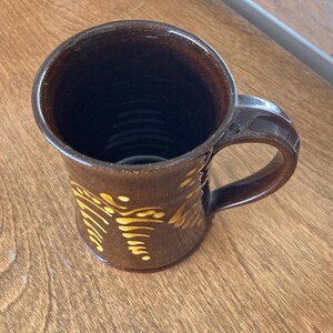 May include: A brown ceramic mug with a handle and a yellow pattern. The mug has a glossy finish and a dark interior. The mug is likely handmade and suitable for hot drinks.