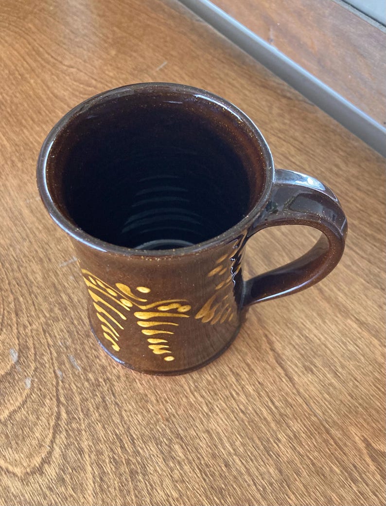 May include: A brown ceramic mug with a handle and a yellow pattern. The mug has a glossy finish and a dark interior. The mug is likely handmade and suitable for hot drinks.