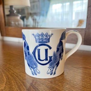 May include: A white ceramic mug featuring a blue design of two horses flanking a central letter U enclosed within a circular border, topped with a crown graphic. The handle is attached to the right side of the cylindrical vessel.