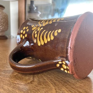 May include: A brown ceramic mug with a gold decorative pattern. The mug features a large handle and a wide base. The gold design consists of dots and curved lines. The mug is resting on a wooden surface.