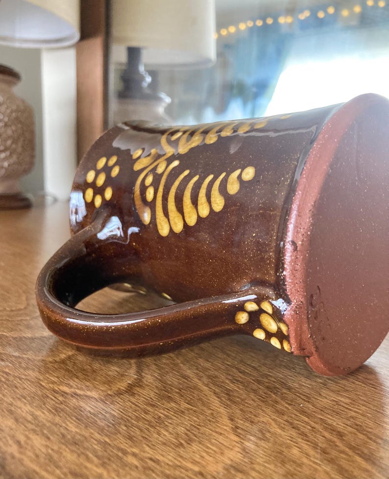 May include: A brown ceramic mug with a gold decorative pattern. The mug features a large handle and a wide base. The gold design consists of dots and curved lines. The mug is resting on a wooden surface.